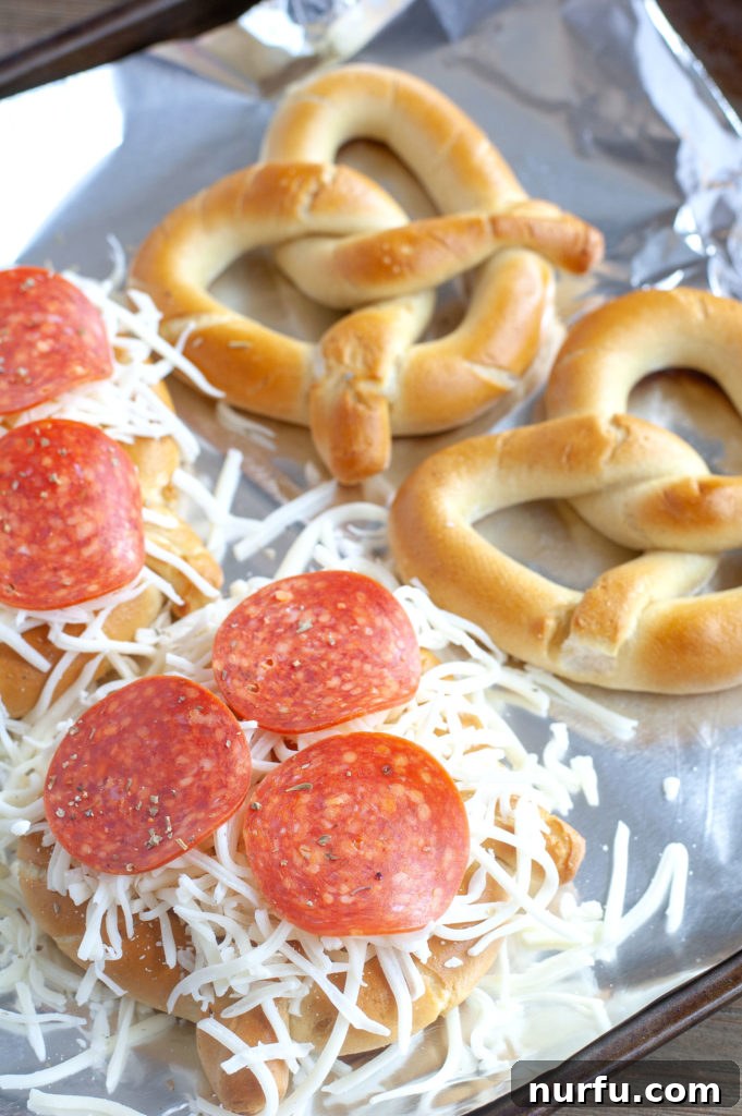 Soft pretzels laid out on a foil-lined baking sheet, topped with shredded cheese and pepperoni, ready for the oven.