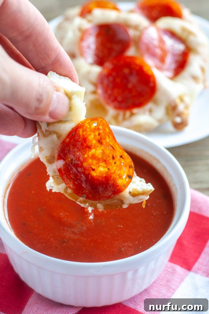A slice of pepperoni pretzel pizza being dipped into a bowl of vibrant red marinara sauce.
