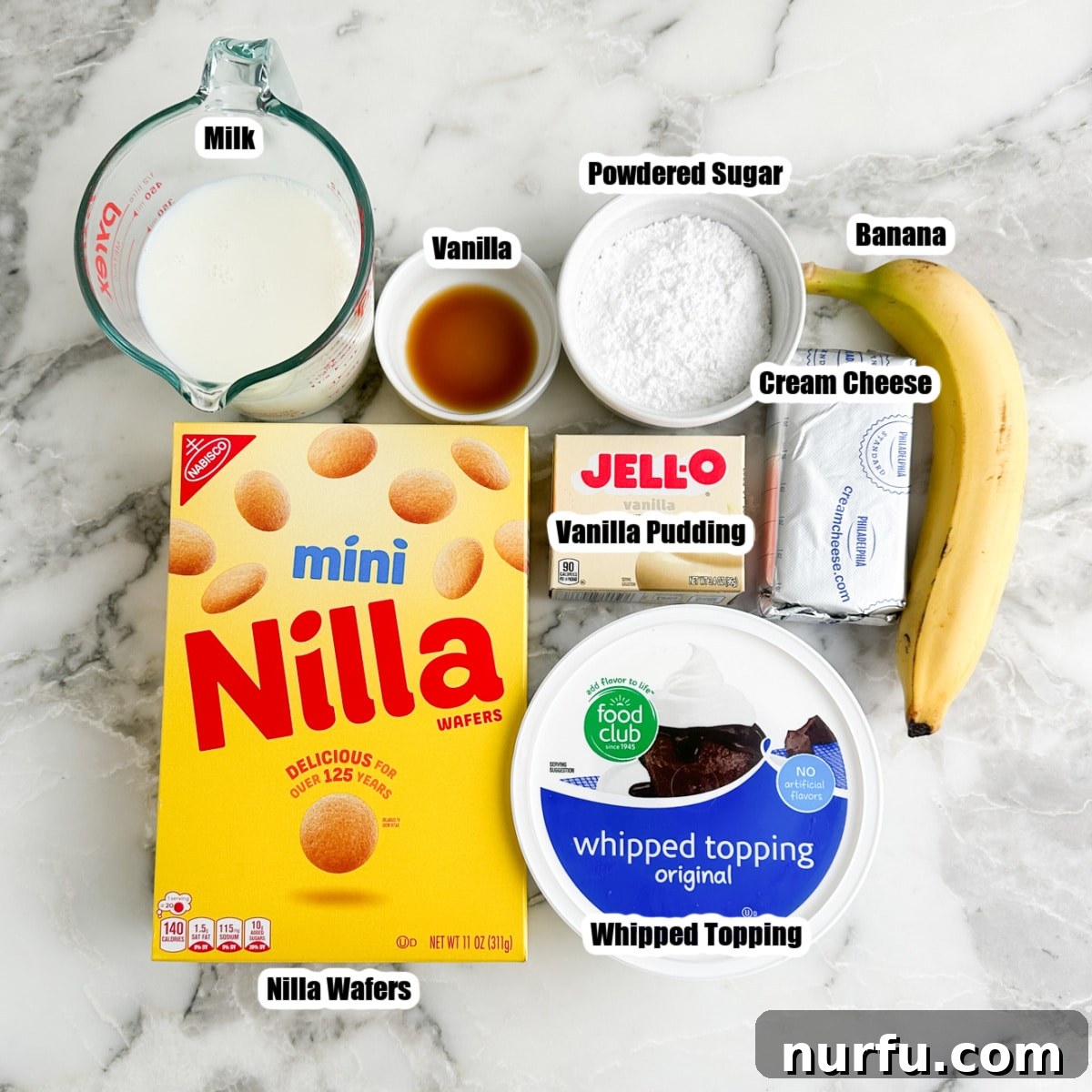 A flat lay photograph showcasing the key ingredients for banana pudding dip: milk, powdered sugar, instant pudding mix, a box of vanilla wafers, whipped topping, and a block of cream cheese.