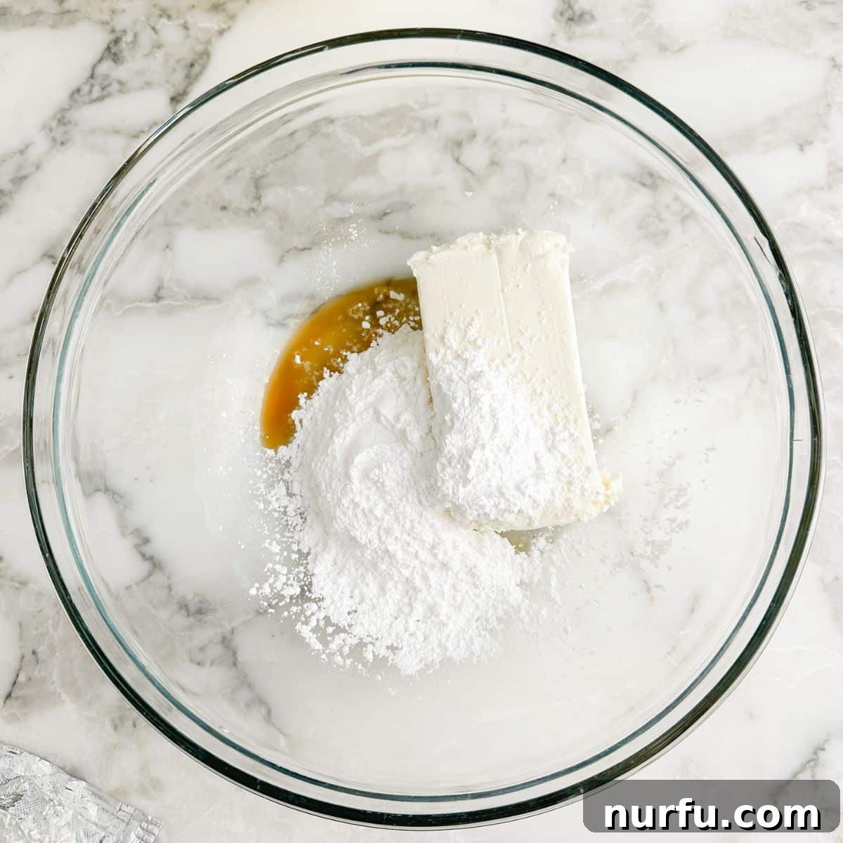 A bowl containing softened cream cheese, powdered sugar, and vanilla extract being beaten with an electric mixer until creamy.