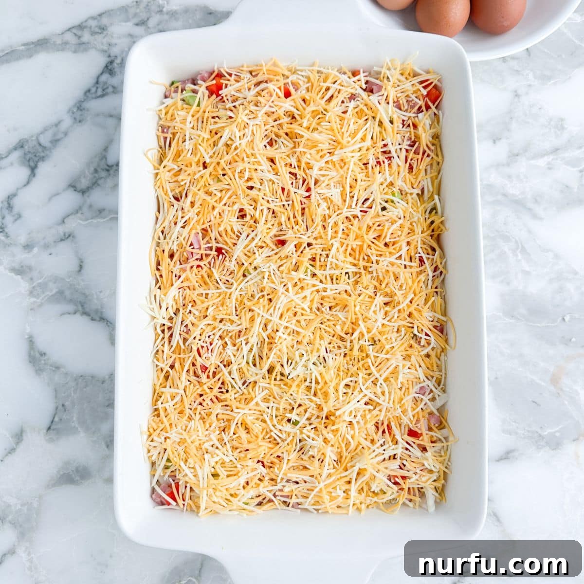 Hearty Farmhouse Casserole 4 A casserole dish with hash browns, topped with colorful diced green onions, red peppers, savory ham, and a generous layer of shredded cheese.