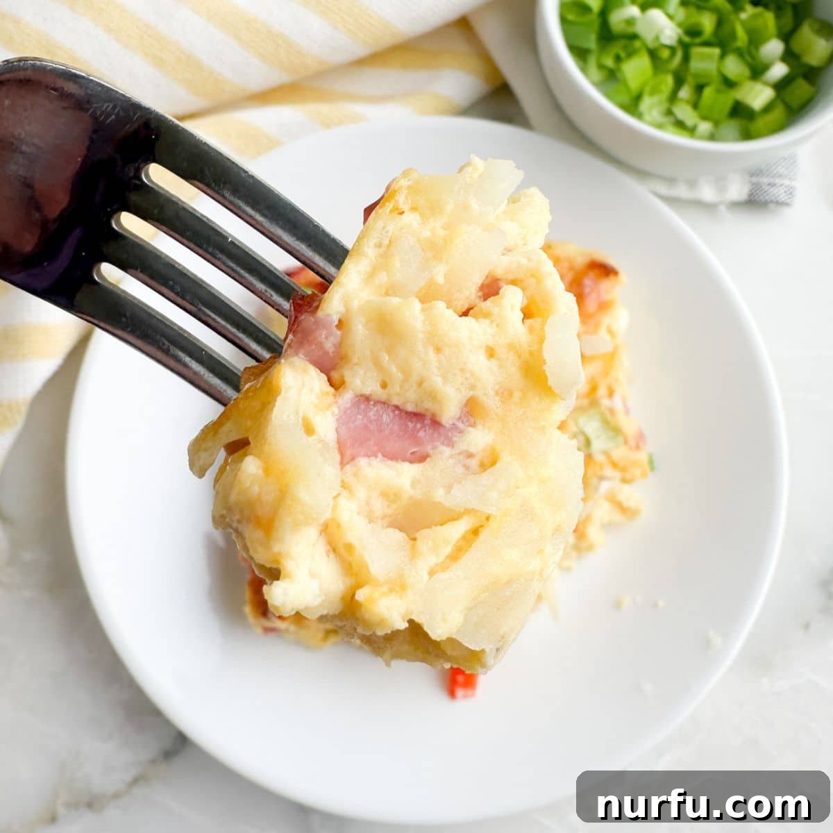 Hearty Farmhouse Casserole 8 A fork piercing into a delicious piece of Farmers Casserole on a white plate, showing the layers of egg, cheese, ham, and hash browns.