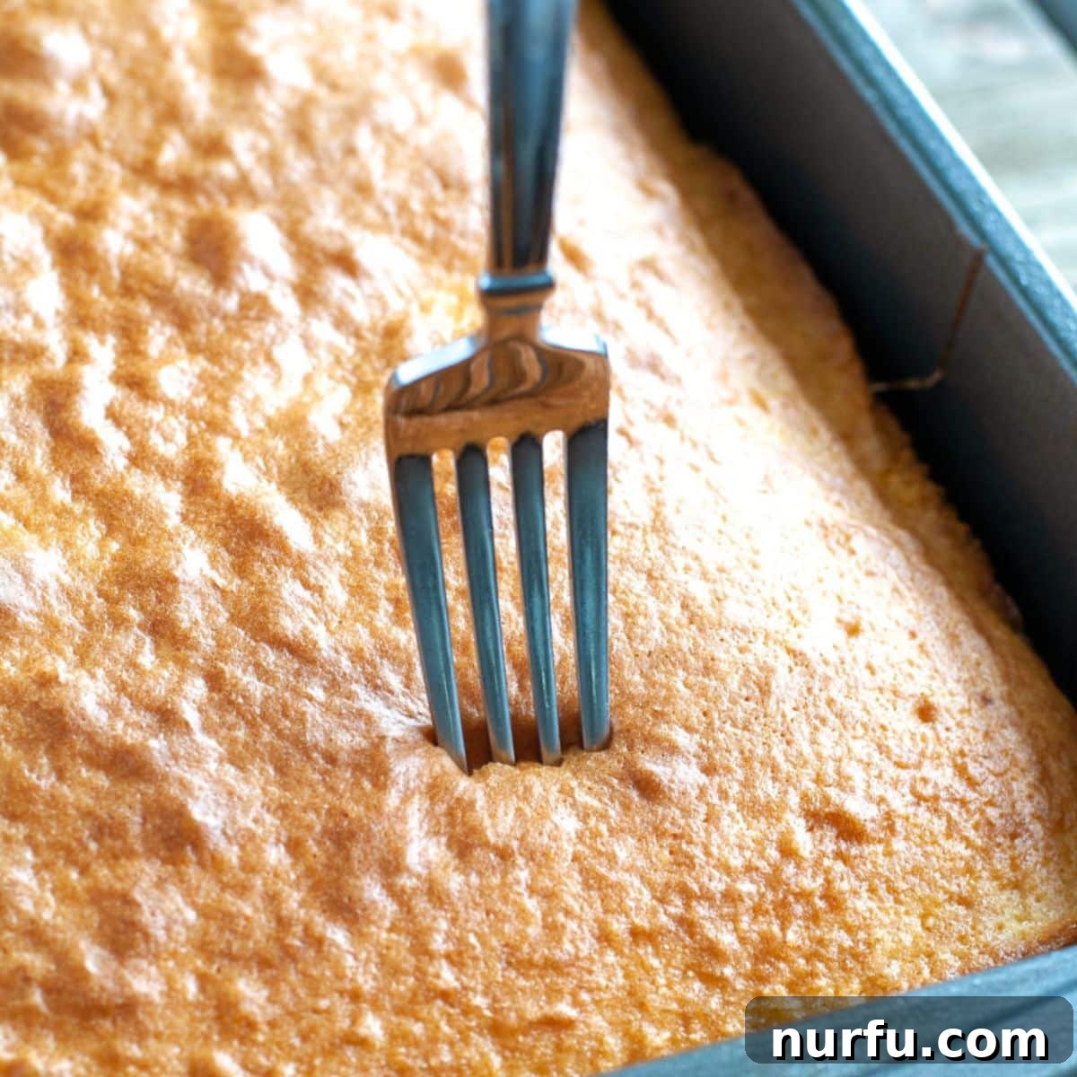 Golden Crunch Butterfinger Poke Cake 3 A freshly baked yellow cake in a 9x13 pan with a fork creating poke holes across its surface.