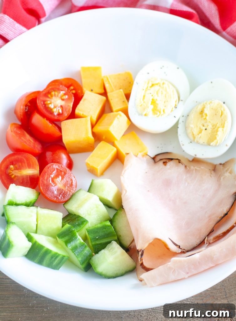 Keto Snack Box A white bowl showcasing individual keto snack box ingredients: tomato halves, cheese cubes, a hard-boiled egg cut in half, cucumber slices, and folded turkey slices.