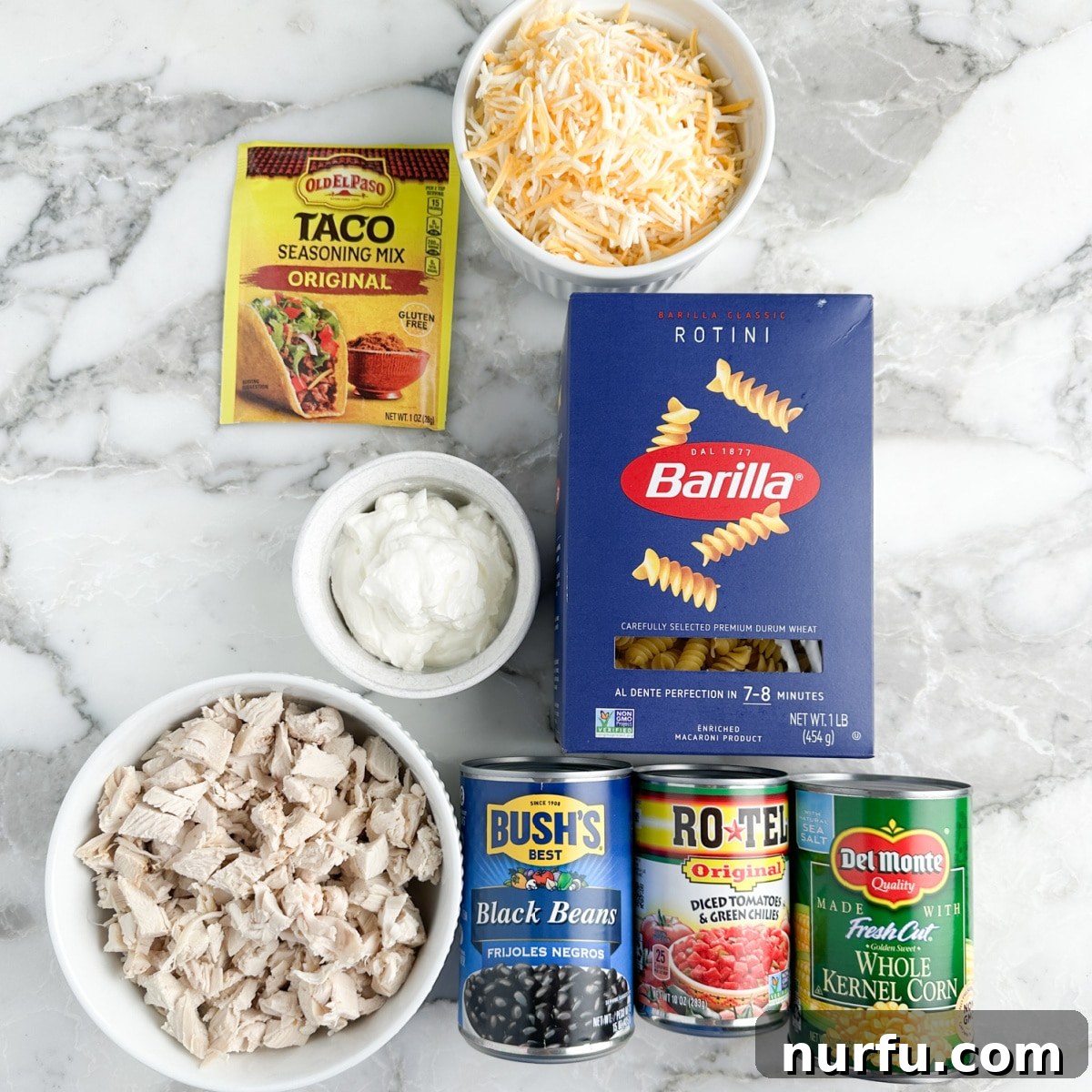 Celebration Chicken Bake 2 Individual ingredients for Fiesta Chicken Casserole including rotini pasta, taco seasonings, shredded cheese, diced chicken, sour cream, cans of beans, tomatoes, and corn.