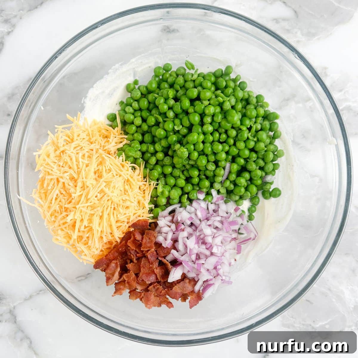 Quick and Creamy Pea Salad 3 A large mixing bowl filled with thawed green peas, shredded cheese, crispy bacon bits, and finely diced red onion, before the creamy dressing is added.