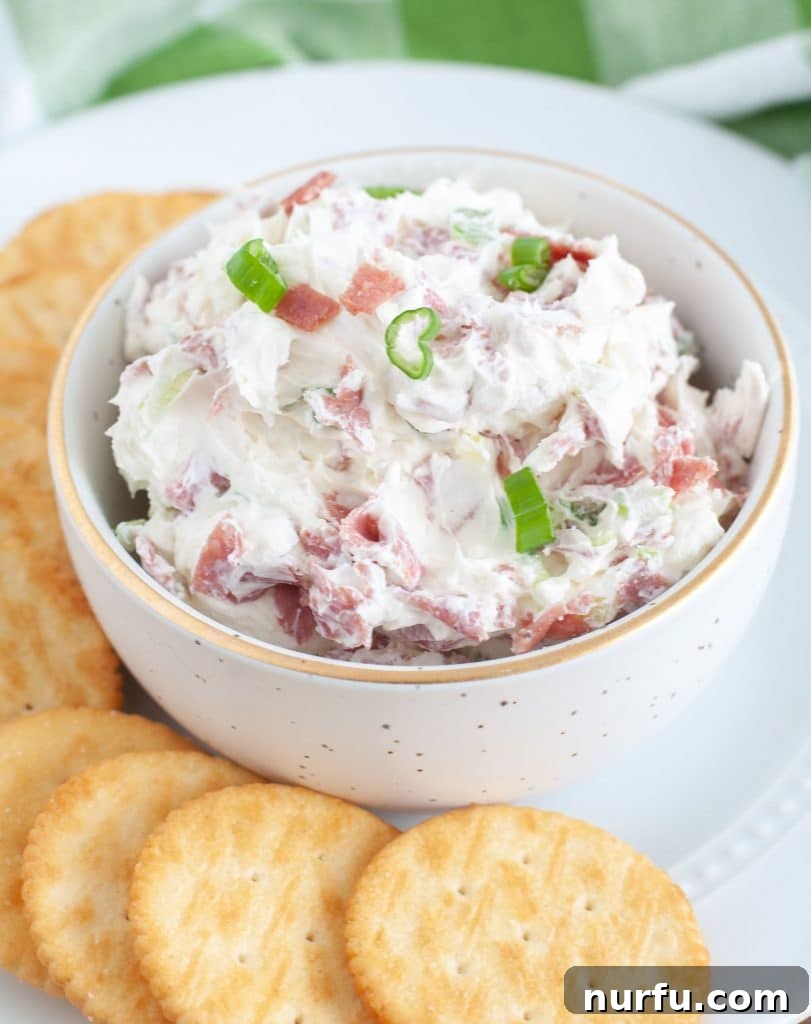 A small white bowl of creamy chipped beef dip, generously garnished with fresh green onions, surrounded by an assortment of crispy crackers on a white serving plate. This savory dip is perfect for parties.