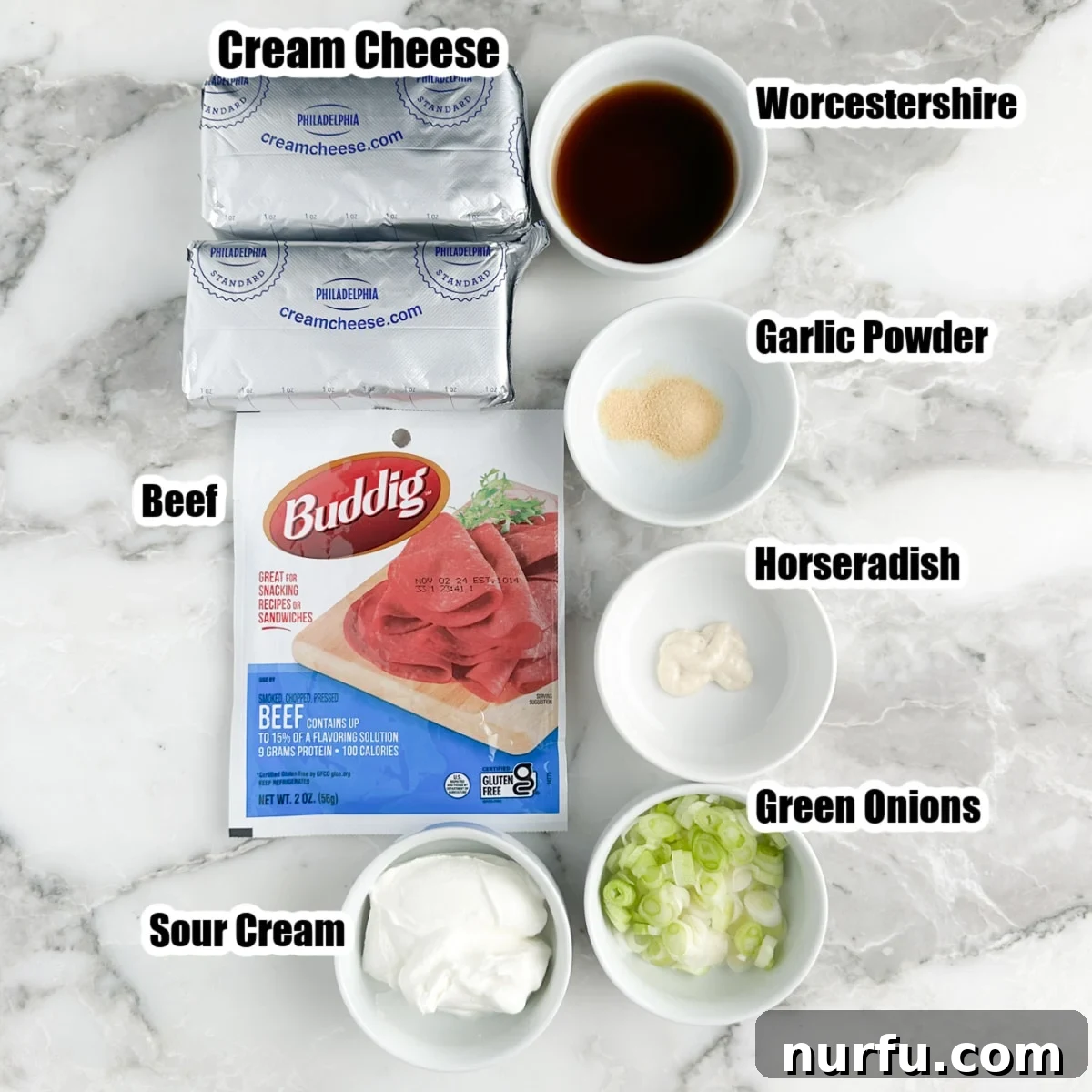 Individual ingredients for Chipped Beef Dip: A package of Buddig dried beef, a block of cream cheese, a small bowl of sour cream, fresh green onions, and a bottle of Worcestershire sauce are neatly arranged on a counter.