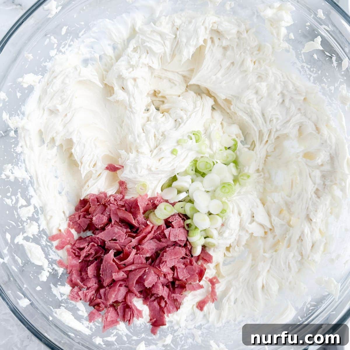A large mixing bowl filled with the creamy chipped beef dip base, now featuring finely diced green onions and chopped chipped beef stirred in, showcasing the vibrant colors and textures before final mixing.