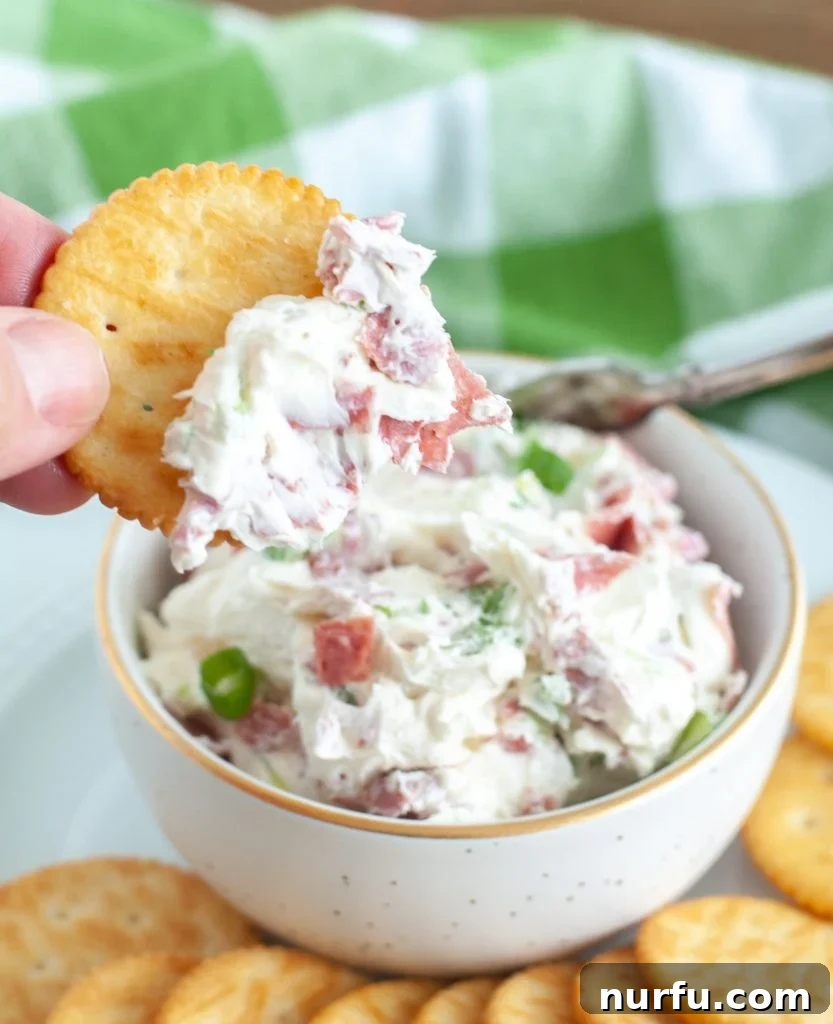 A cracker, generously topped with creamy Chipped Beef Dip and a sprinkle of green onion, is held above the main bowl of dip, ready to be enjoyed. The focus is on the delicious texture and presentation.
