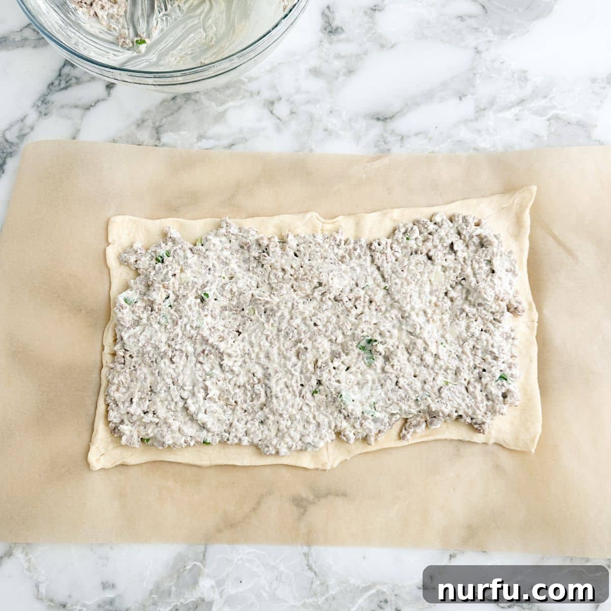 Savory Sausage Spirals 5 A rectangular sheet of crescent dough, seamlessly joined, with a generous and even layer of the creamy sausage and cream cheese mixture spread across its surface, ready for rolling.