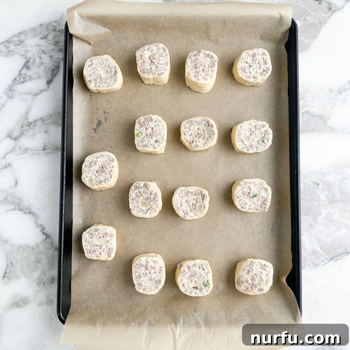 Savory Sausage Spirals 7 Unbaked, thinly sliced sausage pinwheels neatly arranged on a parchment-lined baking sheet, ready for the oven.