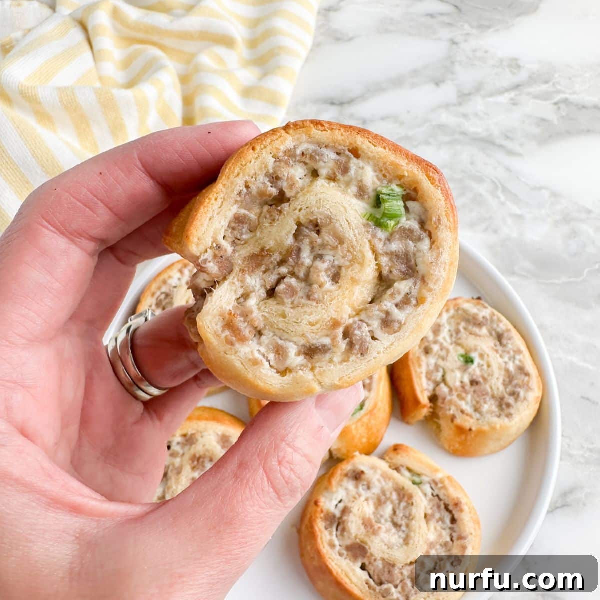 Savory Sausage Spirals 9 A hand holds up a single, perfectly golden-brown sausage pinwheel, showcasing its flaky crescent crust and savory filling, with more pinwheels blurred in the background.