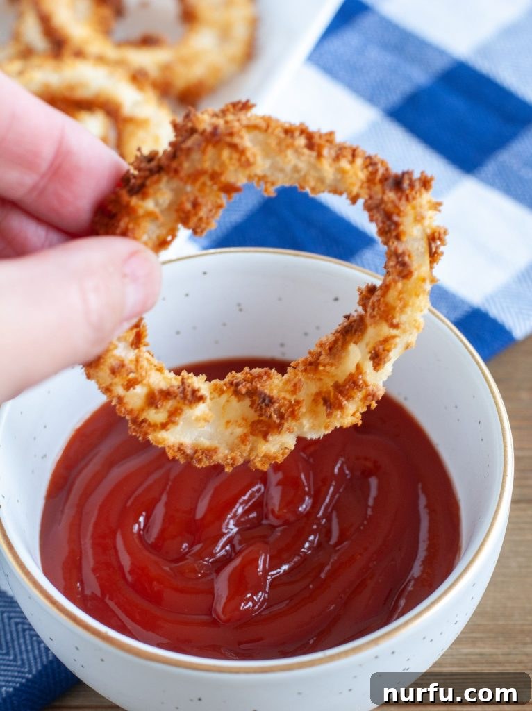 Crispy Air-Fried Onion Rings 5 A perfectly cooked air fryer onion ring being dipped into a small dollop of vibrant red ketchup on a white plate.