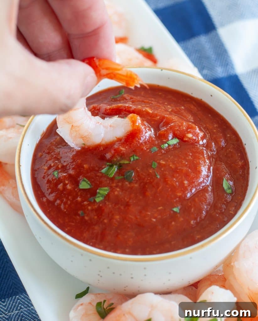 Close-up shot of a cooked shrimp being dipped into a bowl of vibrant homemade cocktail sauce, highlighting the texture and color.