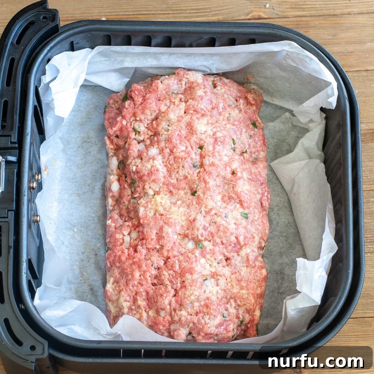 An uncooked meatloaf, shaped and placed on parchment paper, resting inside an air fryer basket, ready for cooking.