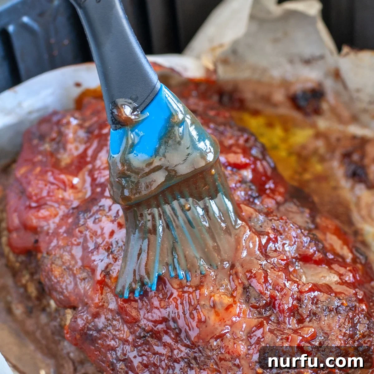 A close-up shot of a partially cooked air fryer meatloaf, gleaming with a fresh application of savory sauce being brushed onto its surface.