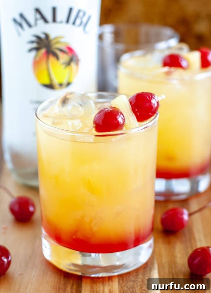 Rum cocktail in a glass with cherries and bottle in background, showcasing the beautiful layered effect of a Malibu Sunset.