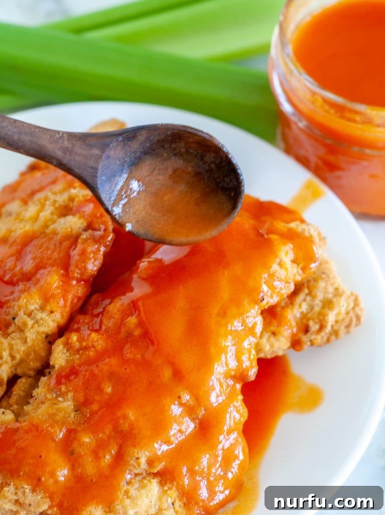 Easy Homemade Buffalo Sauce 6 A spoon overflowing with rich, red homemade buffalo sauce, with pieces of cooked chicken in the background, illustrating a delicious serving suggestion.