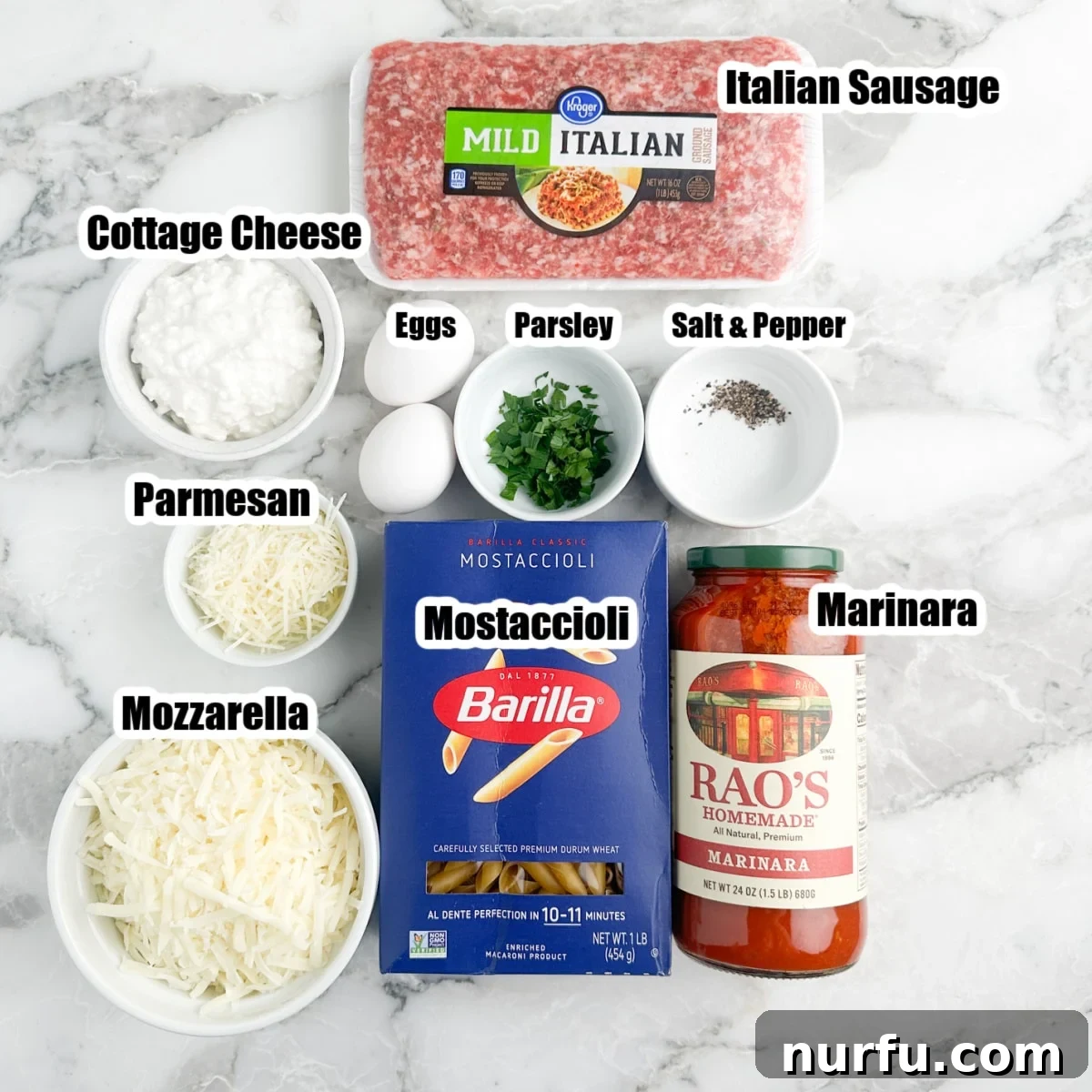 Cheesy Baked Mostaccioli 3 A vibrant flat lay of ingredients for baked mostaccioli: Italian sausage, a bowl of shredded mozzarella, cottage cheese, dried mostaccioli pasta, and a jar of marinara sauce.
