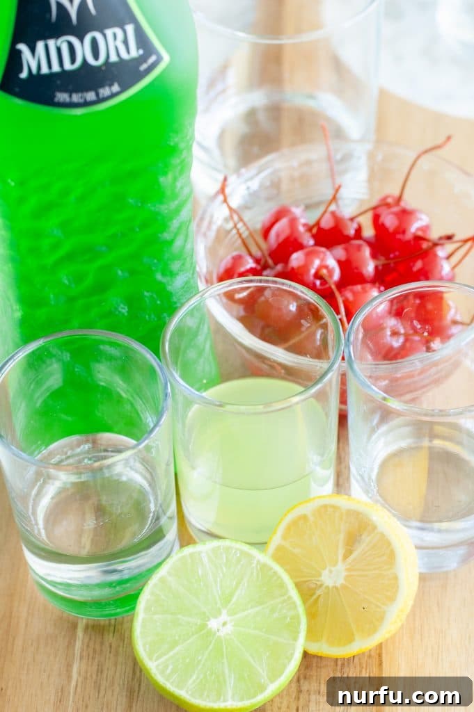 A collection of essential Midori Sour ingredients neatly arranged on a table, including a bottle of Midori, a bowl overflowing with bright red maraschino cherries, fresh yellow lemons, vibrant green limes, and a glass containing clear vodka.