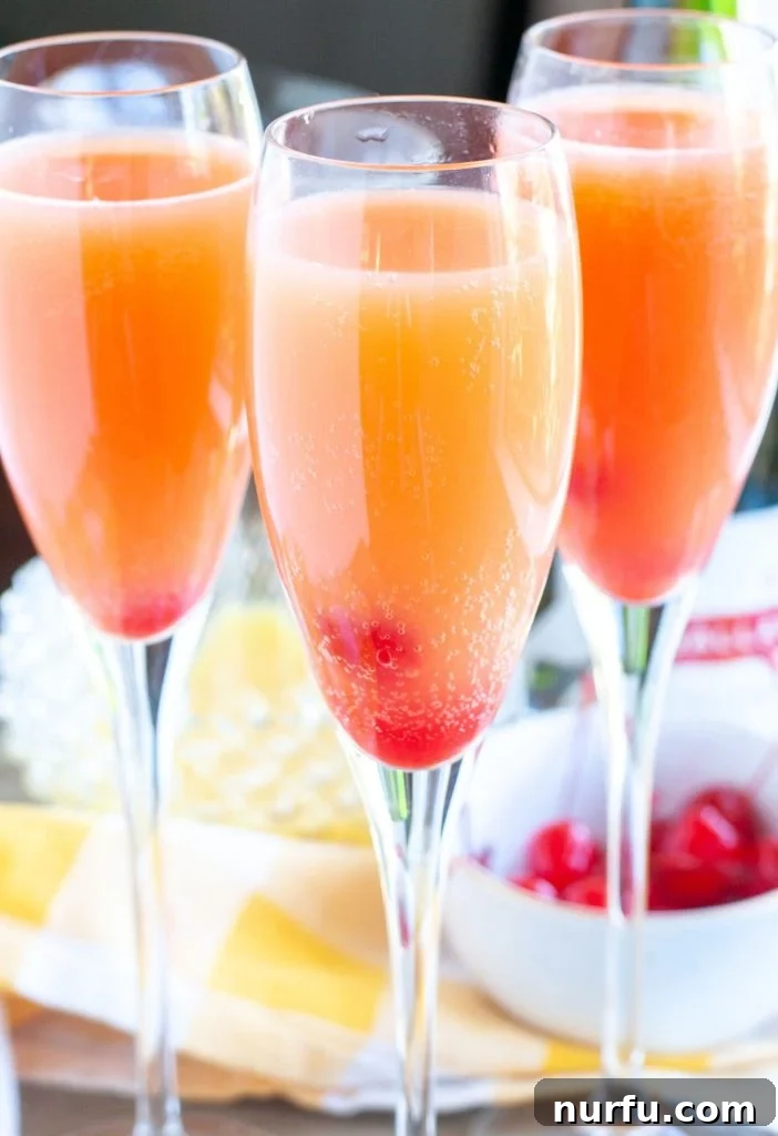 Three glasses of blushing mimosas, garnished with maraschino cherries, on a light background.
