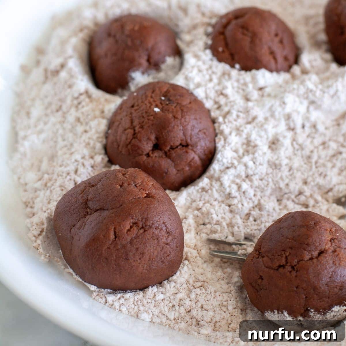 Spiced Chocolate Crinkle Delights 4 Scooped chocolate cookie dough balls being rolled in a bowl of white powdered sugar and cinnamon mixture, ready for baking.