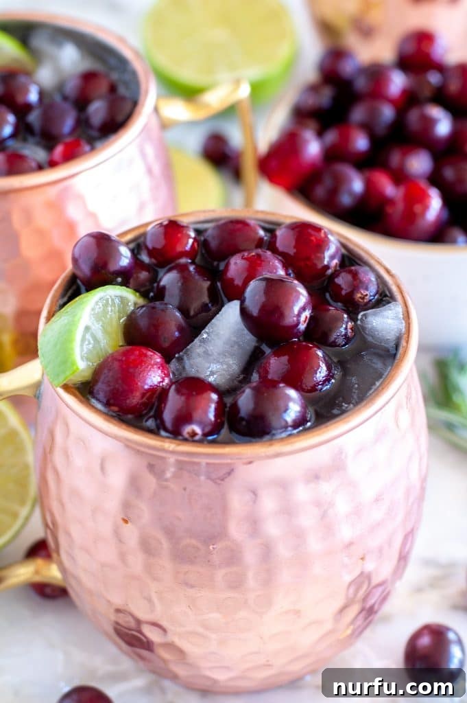 A glistening copper mug brimming with a Cranberry Moscow Mule, garnished with fresh cranberries and a lime wedge, resting on a rustic wooden surface.