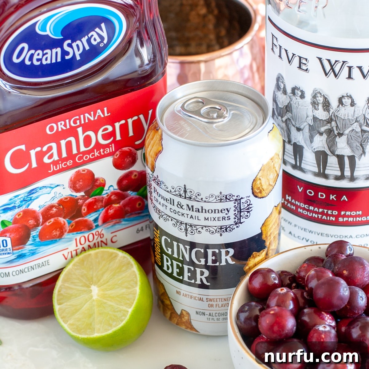 Bottles of premium cranberry juice, spicy ginger beer, and quality vodka arranged together, showcasing the core ingredients for a Cranberry Moscow Mule.