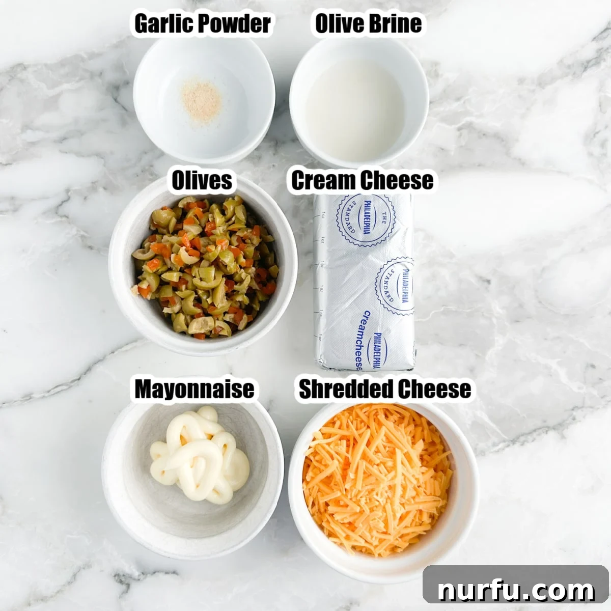 A selection of essential ingredients for the creamy olive dip laid out: a bowl of finely chopped green olives, a block of softened cream cheese, a dollop of creamy mayonnaise, a mound of shredded cheddar cheese, and a small pile of aromatic garlic powder.