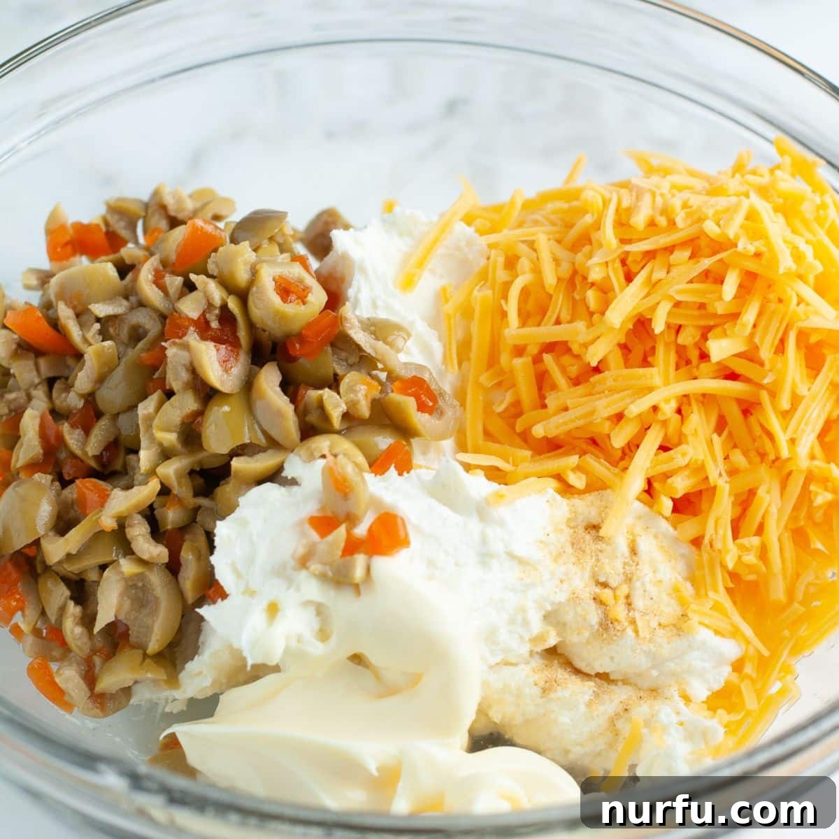 A large mixing bowl containing softened cream cheese, mayonnaise, a pile of finely diced green olives, and a mound of shredded cheddar cheese, perfectly prepped and ready for mixing into a delicious dip.
