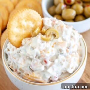 A bowl of creamy green olive dip, garnished with whole olives and fresh herbs, with a cracker dipped into it, ready to be enjoyed as a gourmet appetizer.