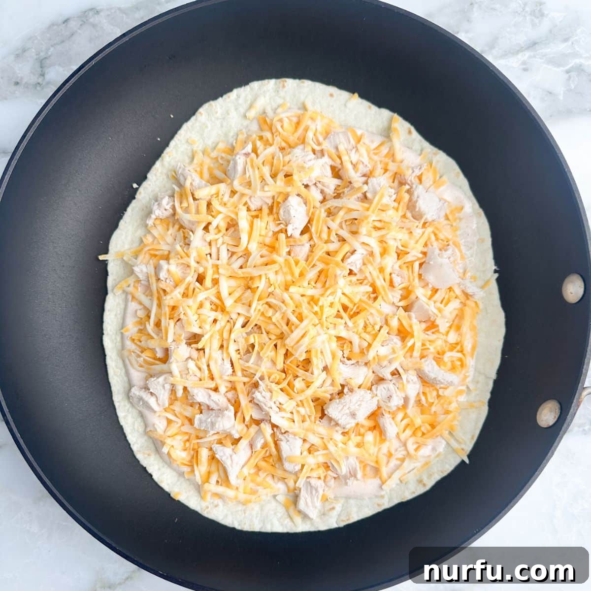 Homemade Taco Bell Chicken Quesadilla Perfection 5 An assembled quesadilla sits in a hot skillet, with a tortilla base topped with a generous layer of shredded cheese and diced chicken, awaiting the top tortilla.