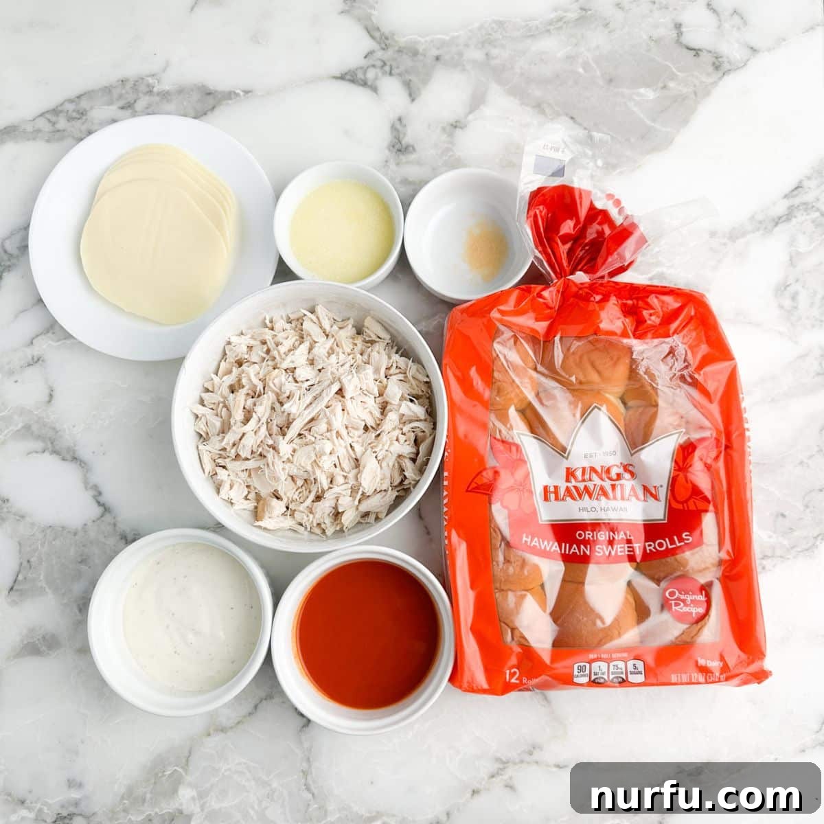 Individual ingredients for buffalo ranch chicken sliders laid out: Hawaiian rolls, bowl of shredded chicken, bowl of buffalo sauce, ranch dressing, and sliced cheese.