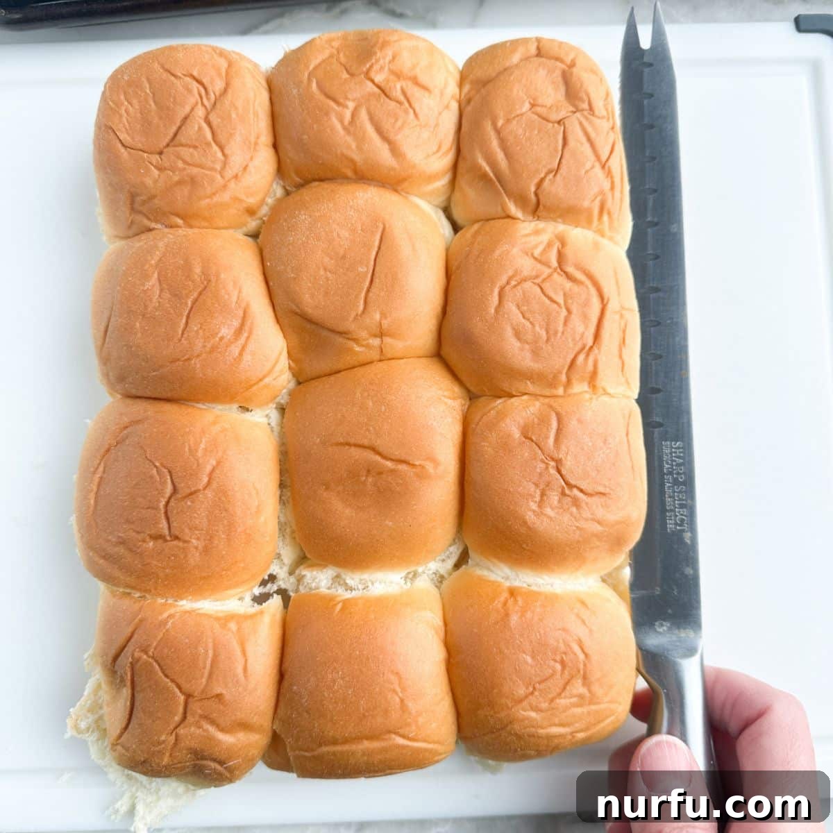 A serrated knife slicing a block of Hawaiian rolls in half horizontally.