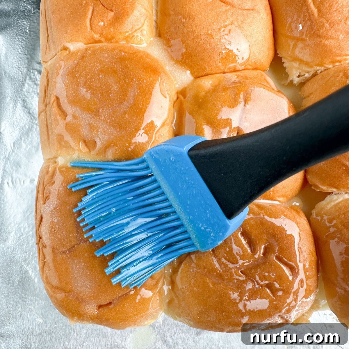 A hand brushing melted garlic butter over the tops of assembled Hawaiian roll sliders before baking.