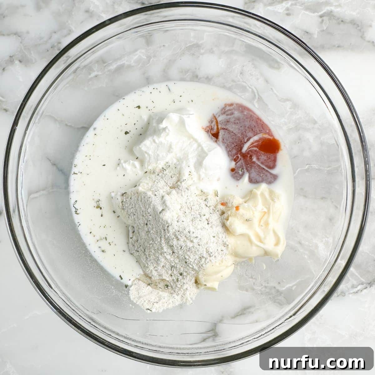 A mixing bowl containing sour cream, mayonnaise, ranch seasoning, milk, and buffalo sauce, ready to be combined.