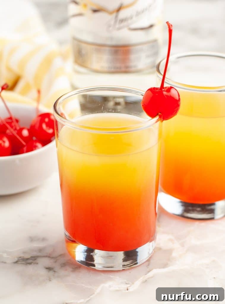 A beautifully layered Pineapple Upside Down Cake Shot, showcasing its orange and red hues, garnished with a maraschino cherry.