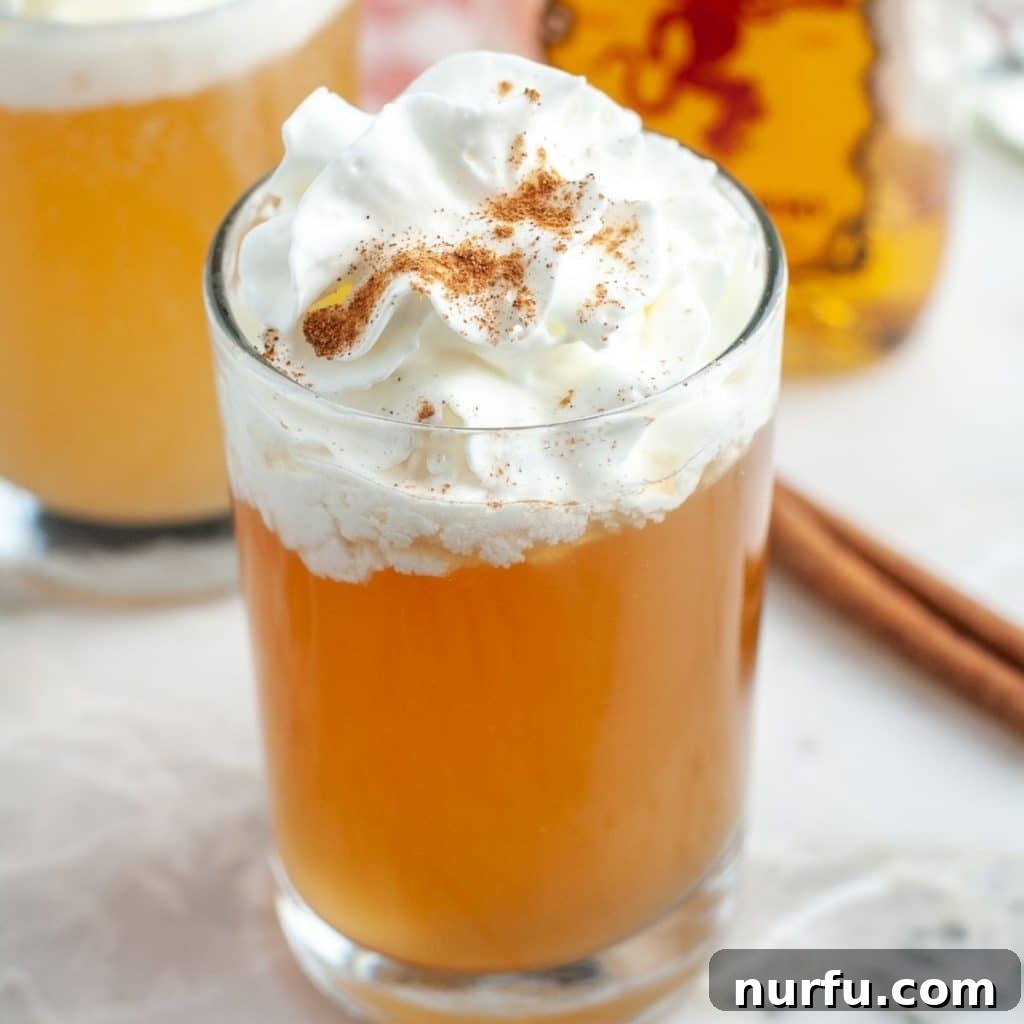 Spiced Apple Pie Shooter 2 A shot glass filled with a golden apple pie cocktail, topped with a generous swirl of whipped cream and a sprinkle of cinnamon spice. The drink perfectly emulates the rich flavors of a classic apple pie.