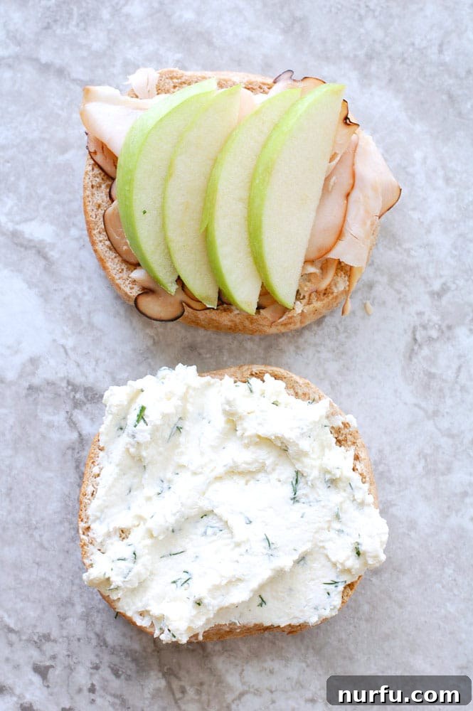 Open Face Turkey Apple Bagel An inviting open-face bagel sandwich, highlighting generous slices of honey turkey and thinly cut green apple, with a smooth, herby cream cheese spread base.