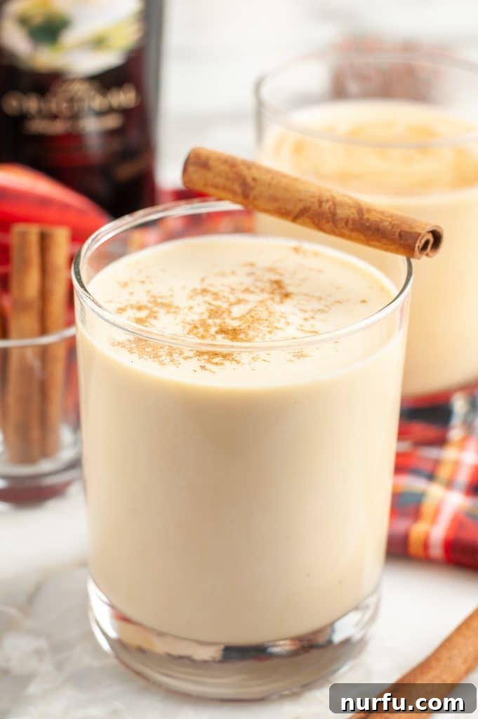 Close-up of a glass of eggnog cocktail, garnished with a cinnamon stick, against a softly blurred festive background.