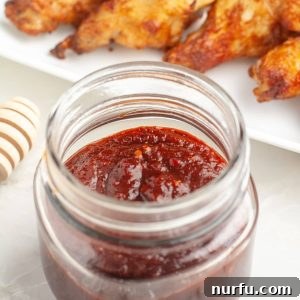Jar of rich, red honey chipotle sauce, with blurred chicken wings in the background, suggesting a delightful meal.