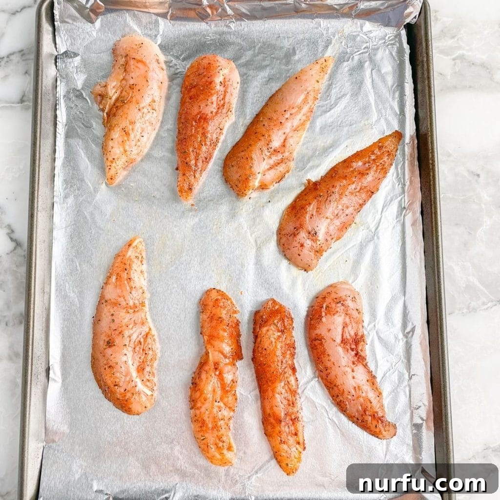 Uncooked, seasoned chicken tenders laid out on a foil-lined baking pan, ready for the oven.