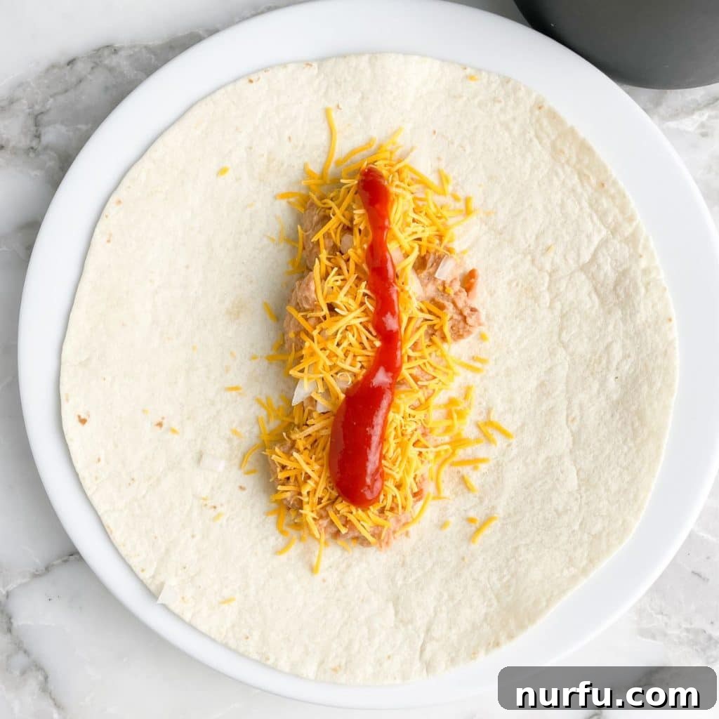 Recreate the Iconic Taco Bell Bean Burrito at Home 5 A partially assembled bean burrito on a tortilla, with refried beans, diced onion, shredded cheese, and red sauce layered on top.