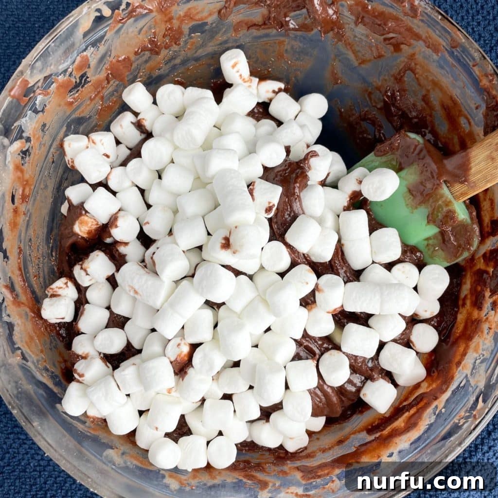 A bowl of melted chocolate fudge, now with mini marshmallows stirred in, ready to be poured into the prepared pan.
