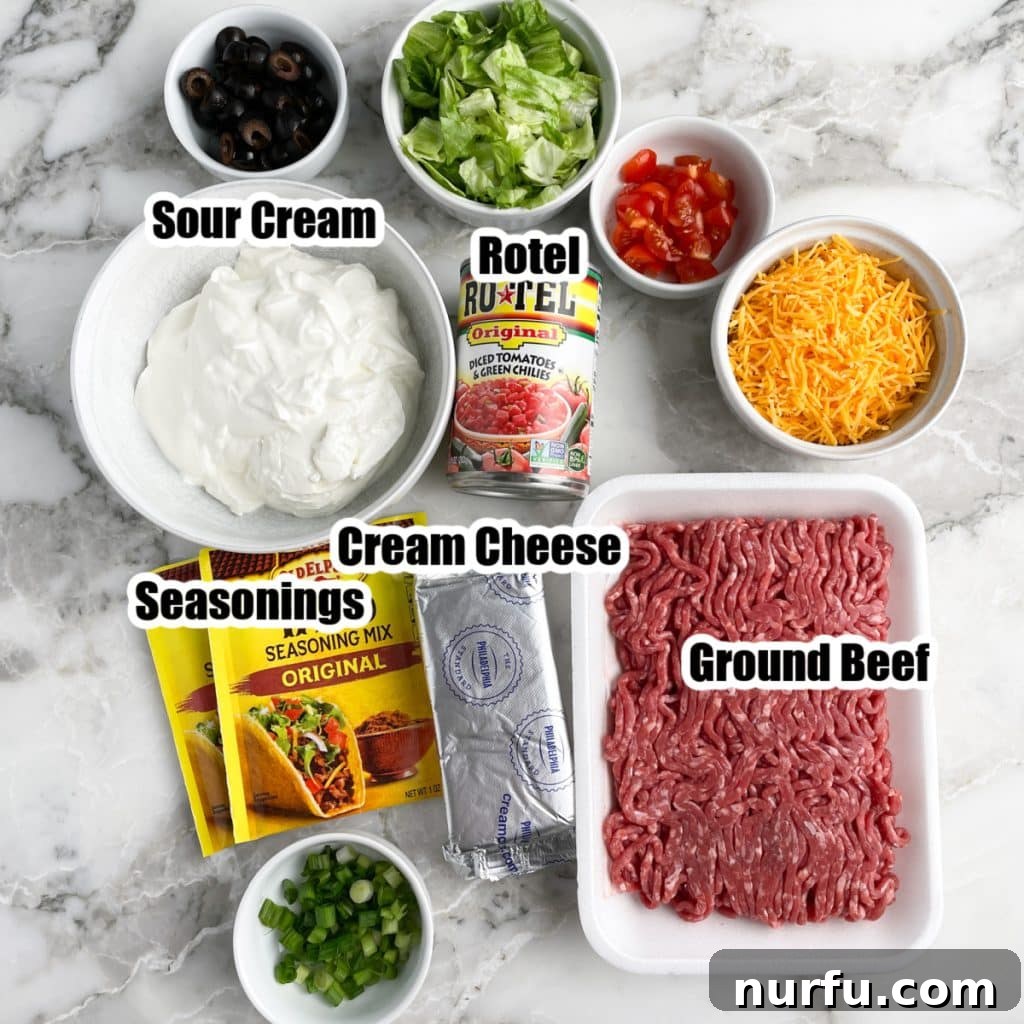 Loaded Ground Beef Taco Fiesta Dip 3 Collection of key ingredients for ground beef taco dip: ground beef, a bowl of sour cream, cream cheese, taco seasoning packets, a bowl of sliced black olives, shredded lettuce, and diced tomatoes.