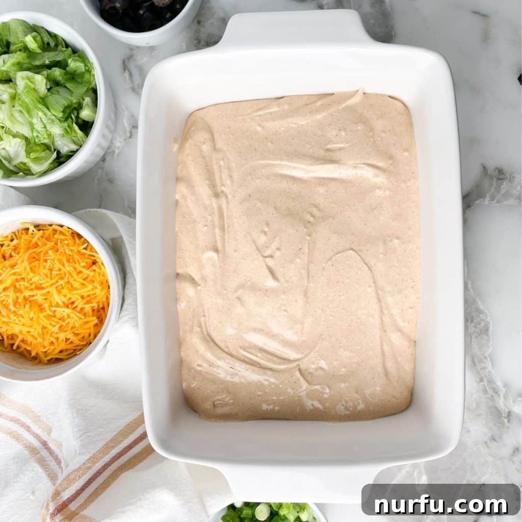 Loaded Ground Beef Taco Fiesta Dip 4 A casserole dish with a creamy mixture, ready to be layered with other ingredients, alongside bowls of shredded lettuce and cheese.