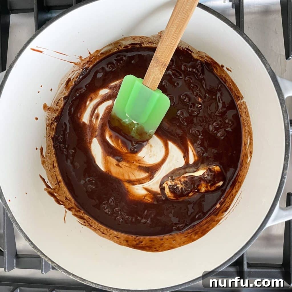 A saucepan on a stovetop with melted butter and chocolate, being stirred by a spatula to combine into a smooth, glossy mixture.