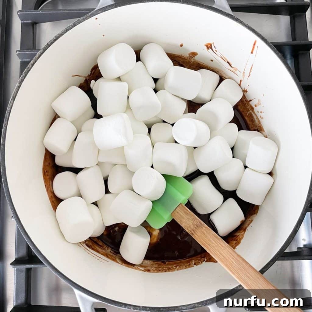 Large marshmallows melting into a rich chocolate and butter mixture in a saucepan, stirred with a spatula, creating a gooey base.