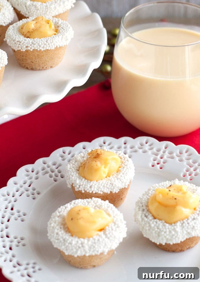 Eggnog cookie cups Festive eggnog cookie cups, beautifully garnished with sprinkles and a dusting of nutmeg, are arranged next to a glass of creamy eggnog, setting a perfect holiday scene.